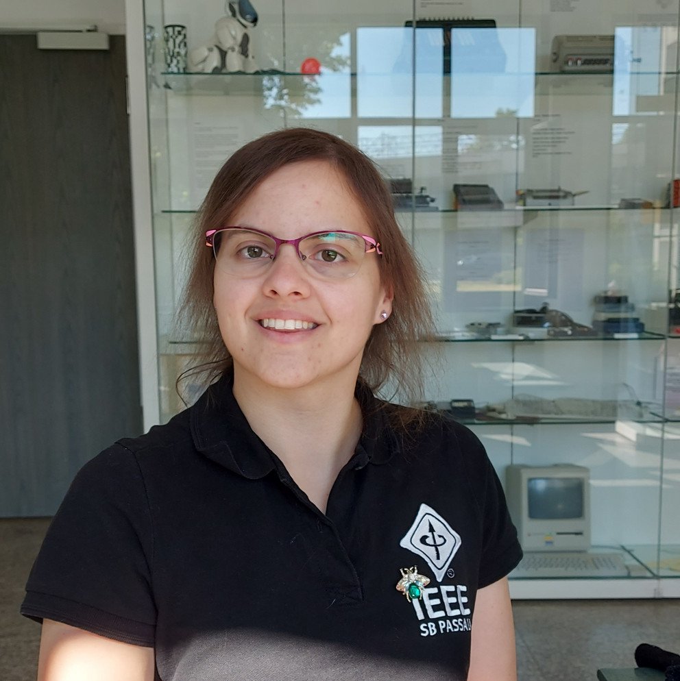 This image shows Marion Christl. She has brown hair, wears glasses with a colorful frame and wears a poloshirt with the IEEE logo of the IEEE student branch Passau. She is sitting in the Mathematics museum in the FIM.