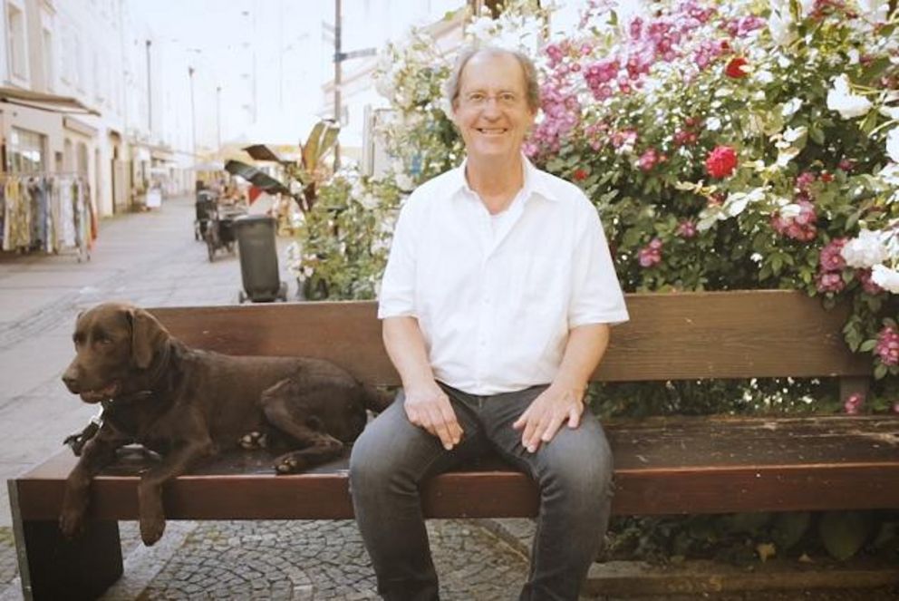 Illustration zur TRIO-Aktion "Science Bench" mit Prof. Dr. Herrmann de Meer.  Er sitzt freundlich lächelnd auf einer Bank in der Passauer Innenstadt, neben ihm ein brauner Hund.